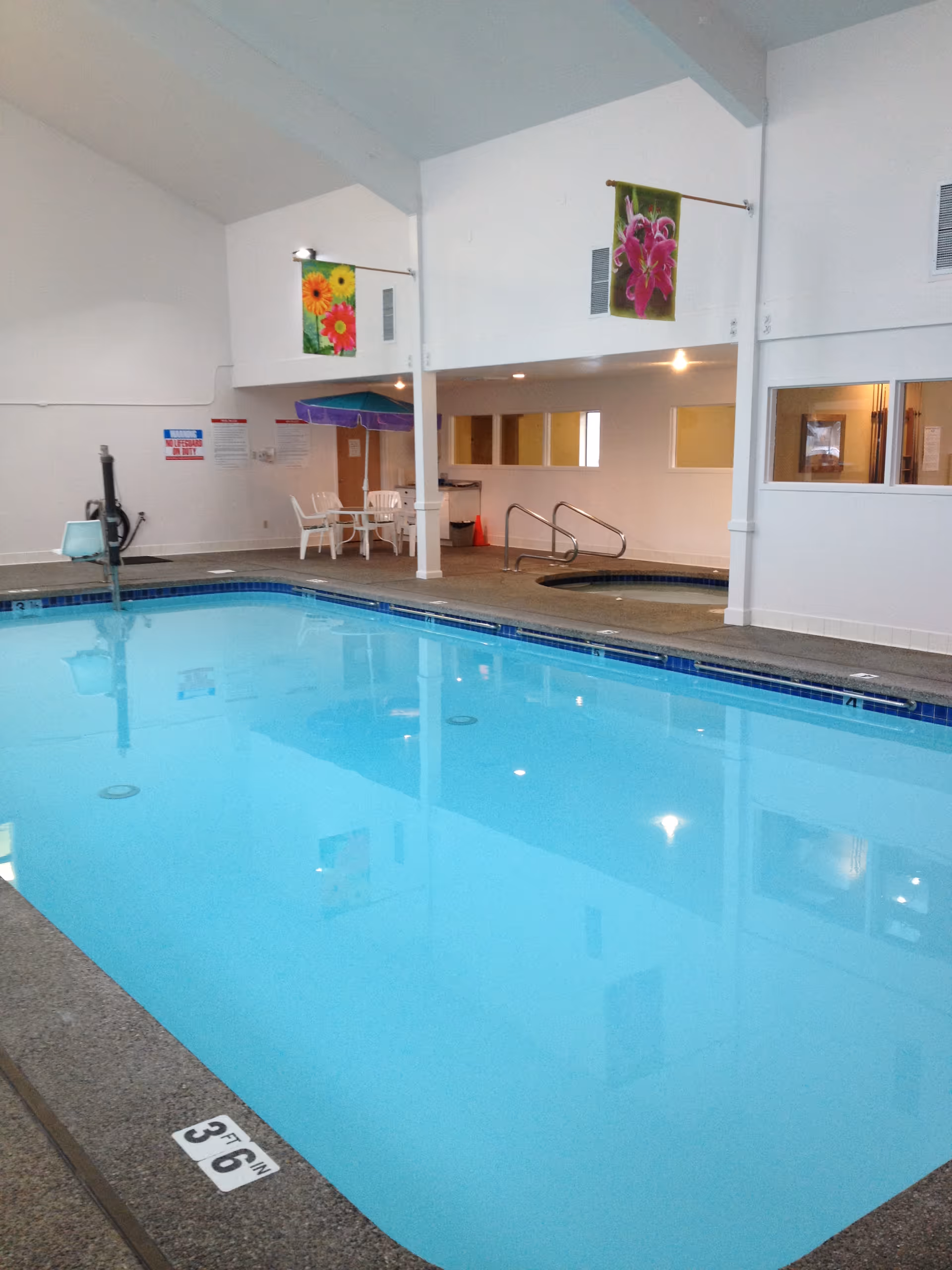 Indoor swimming pool with clear blue water, adjacent hot tub, white walls, and ceiling. There are colorful flower banners hanging on the walls, white plastic chairs and tables with a purple umbrella in the corner, and safety signs on the wall.
