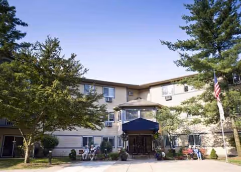 Exterior view of American House Westland Joy with entrance, flag, and trees; guests sit outside.