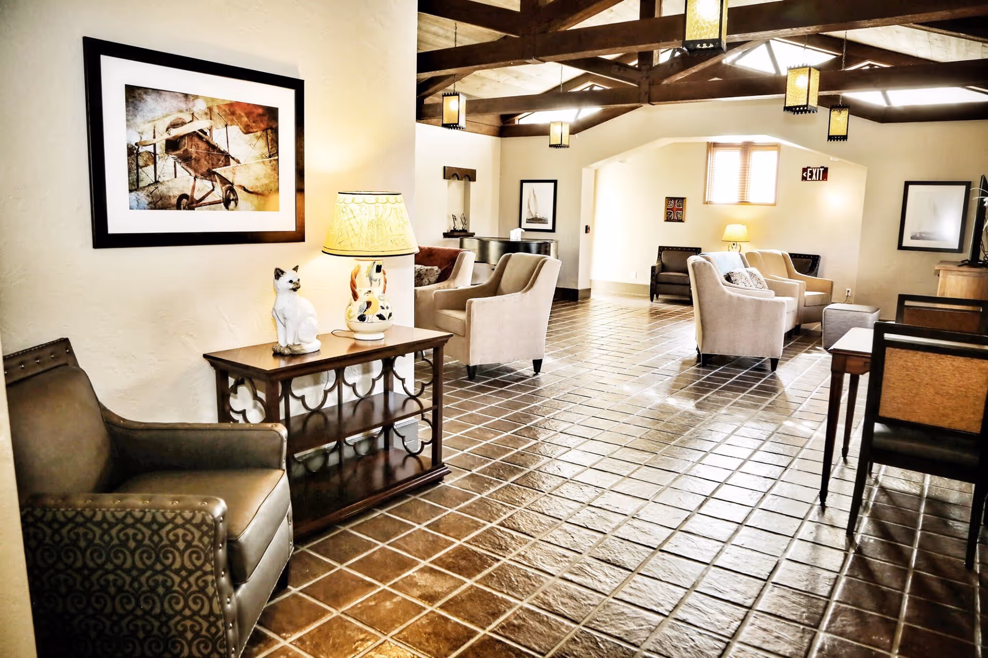 A spacious living room area with tiled flooring and exposed wooden beams on the ceiling. The room is furnished with multiple armchairs, a side table with a lamp and a ceramic cat figurine, framed artwork on the walls, and hanging lantern-style lights. There is an exit sign above a window in the background.
