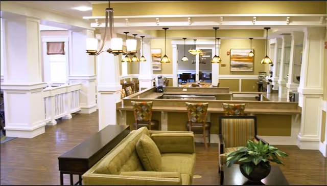 Interior view of a senior living facility lounge area with a green sofa, patterned chairs, a coffee table with a potted plant, and a large counter with bar stools. The room features warm lighting with multiple hanging pendant lights and framed artwork on the walls.
