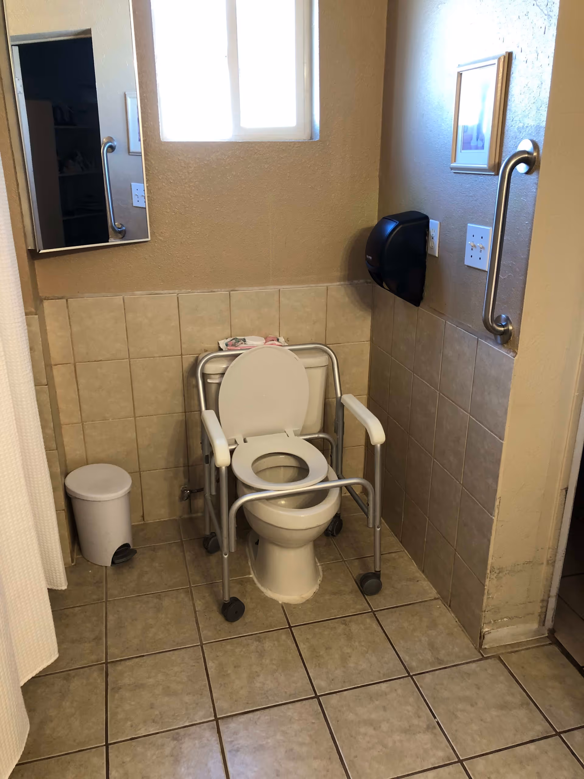 A bathroom with beige tiled floor and walls halfway up. There is a white toilet with a raised toilet seat and arm supports on wheels placed over it. A small white trash can is next to the toilet. A mirror is mounted on the left wall, and a black paper towel dispenser is mounted on the right wall. There is a grab bar on the right wall and a small window above the toilet letting in light.