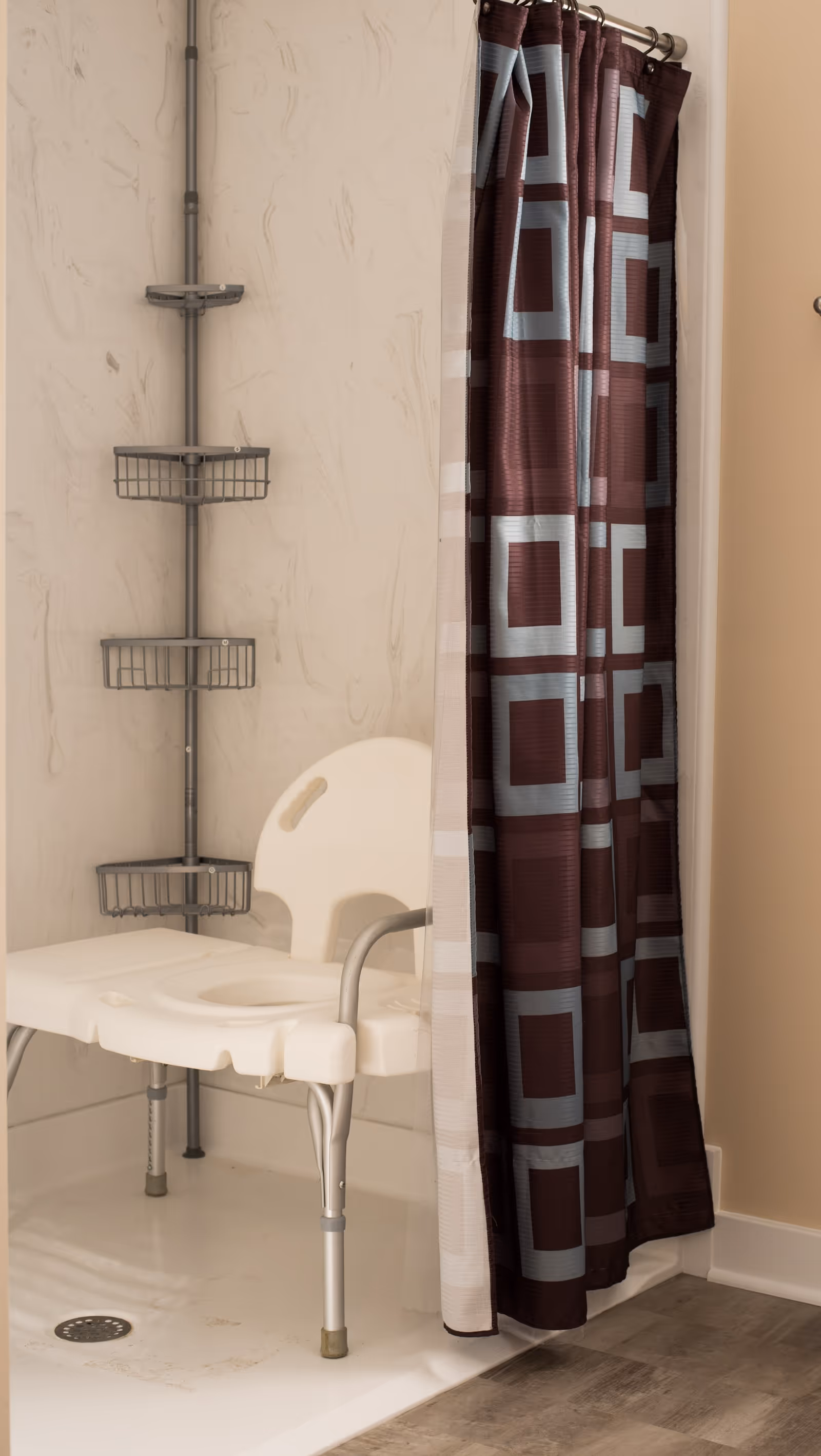 A shower area with a white shower chair and a vertical metal rack with three baskets for toiletries. The shower curtain is partially closed and features a geometric pattern with squares in shades of brown and blue. The floor outside the shower is wood-patterned.