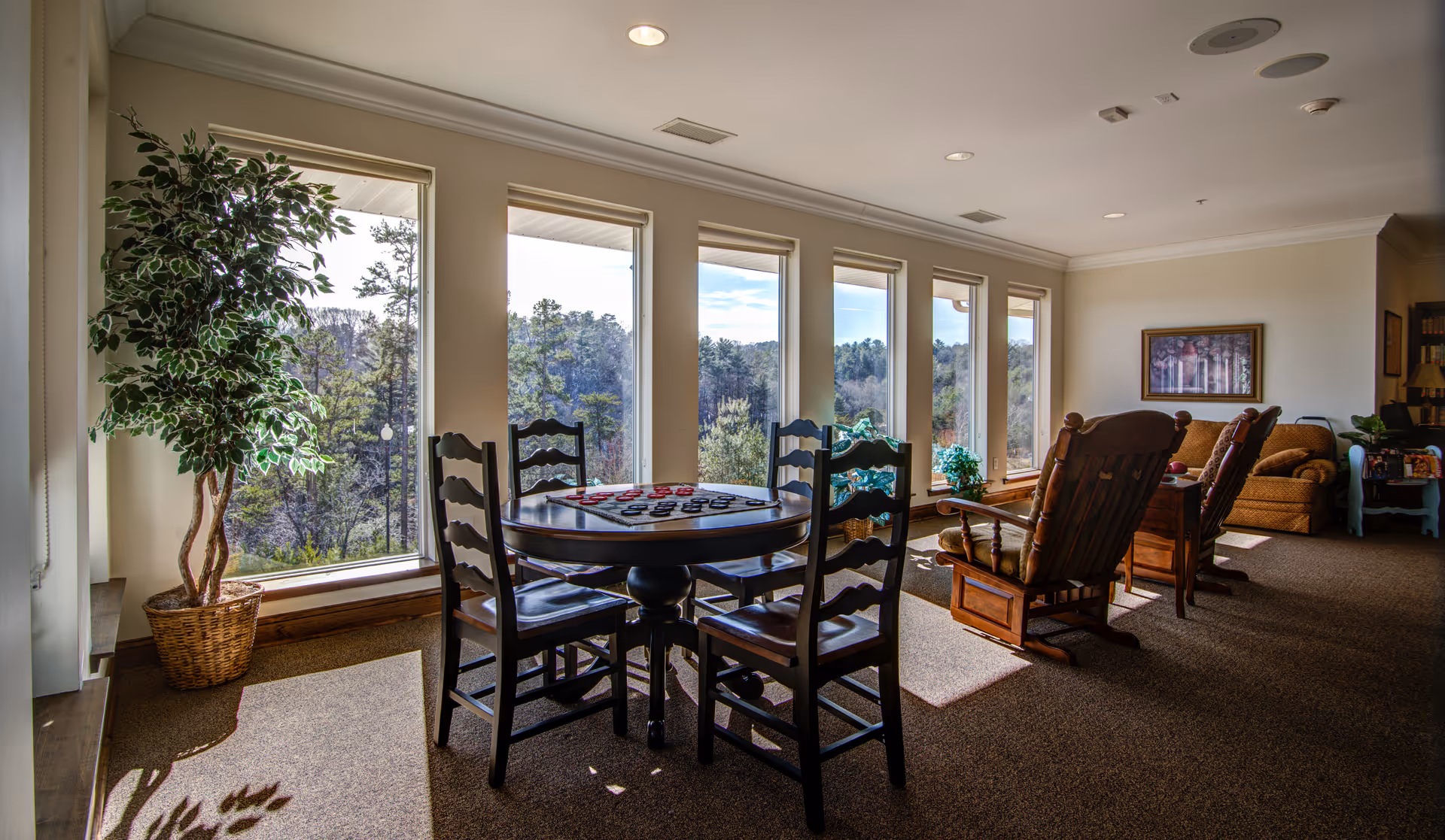Sunlit communal sitting area with a round game table and chairs, wooden recliners and large windows overlooking trees.