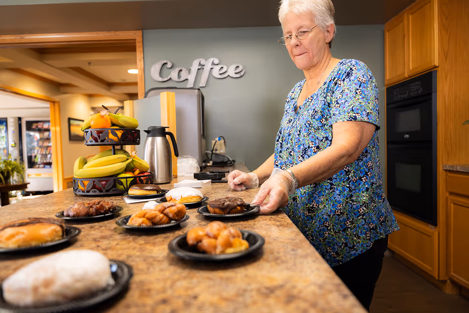 An elderly woman wearing a blue floral shirt and gloves arranges plates of assorted pastries on a kitchen counter. Behind her, there is a tiered fruit stand with bananas and oranges, a coffee pot, and a wall sign that says 'Coffee'. The setting appears to be a warm, well-lit kitchen area.