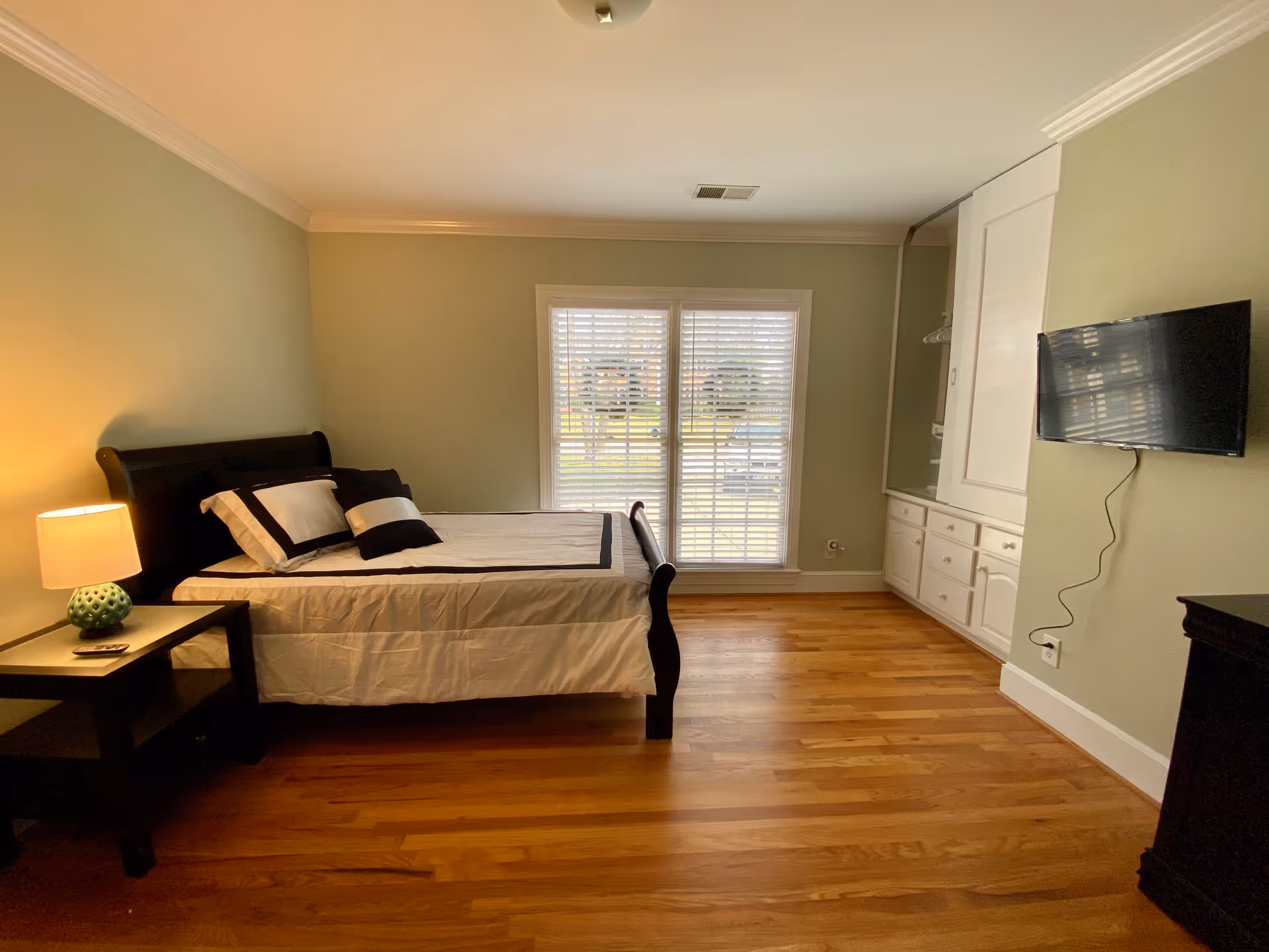 A bedroom with a wooden floor, a bed with white and black bedding, a nightstand with a lamp, a wall-mounted TV, and a large window with white blinds letting in natural light. There is also a built-in cabinet with drawers and a mirror on the right side of the room.