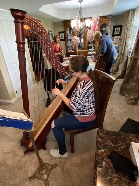 A woman seated on a wooden chair playing a large wooden harp in a carpeted room. In the background, two people are standing near a dining table decorated with balloons and various items. The room has wooden furniture, including a china cabinet and curtains on the windows.