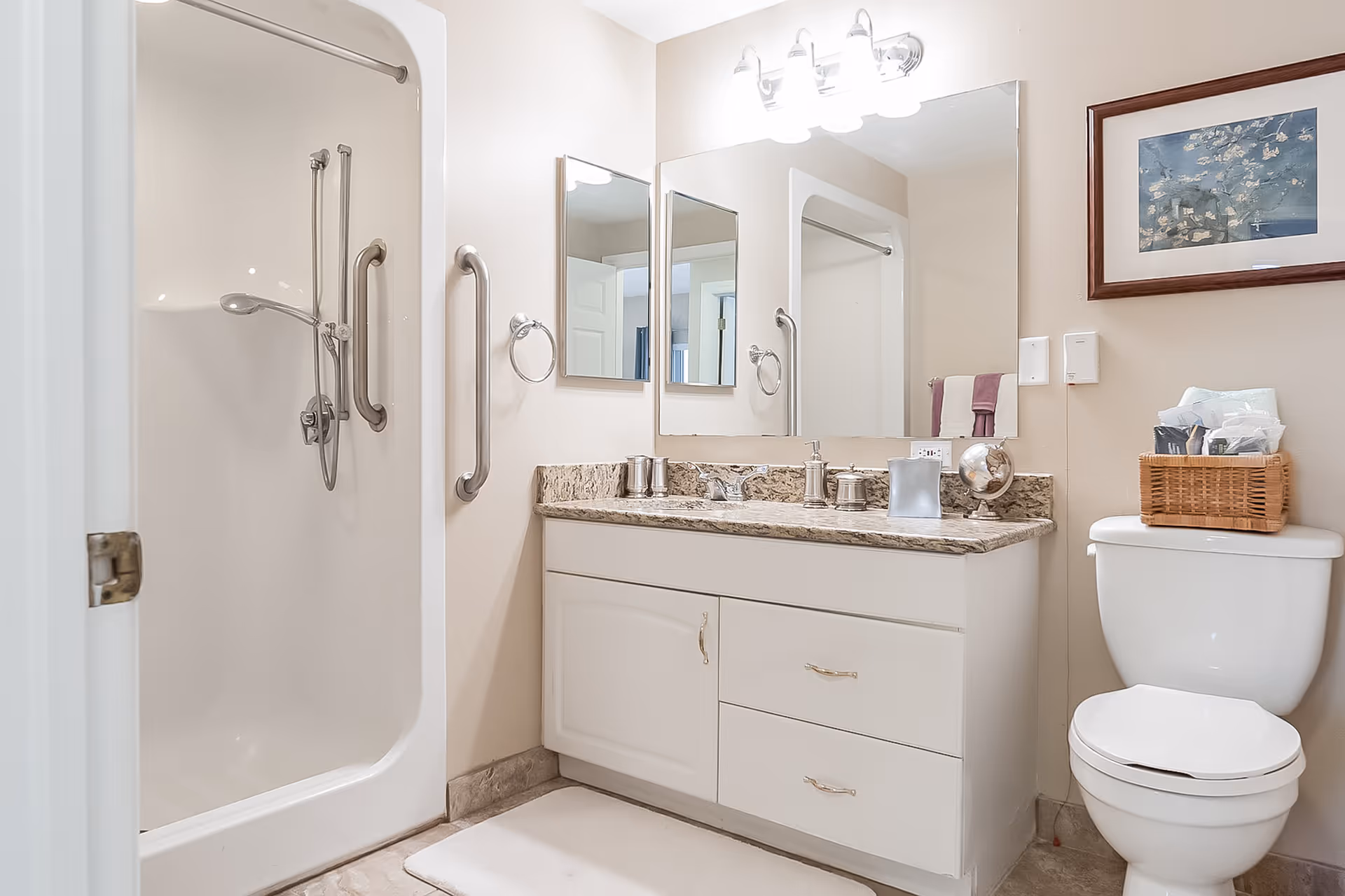 A clean and well-lit bathroom featuring a white shower with grab bars, a vanity with a granite countertop, a large mirror, and a toilet with a wicker basket on top. There is a framed picture on the wall above the toilet and a white bath mat on the floor.