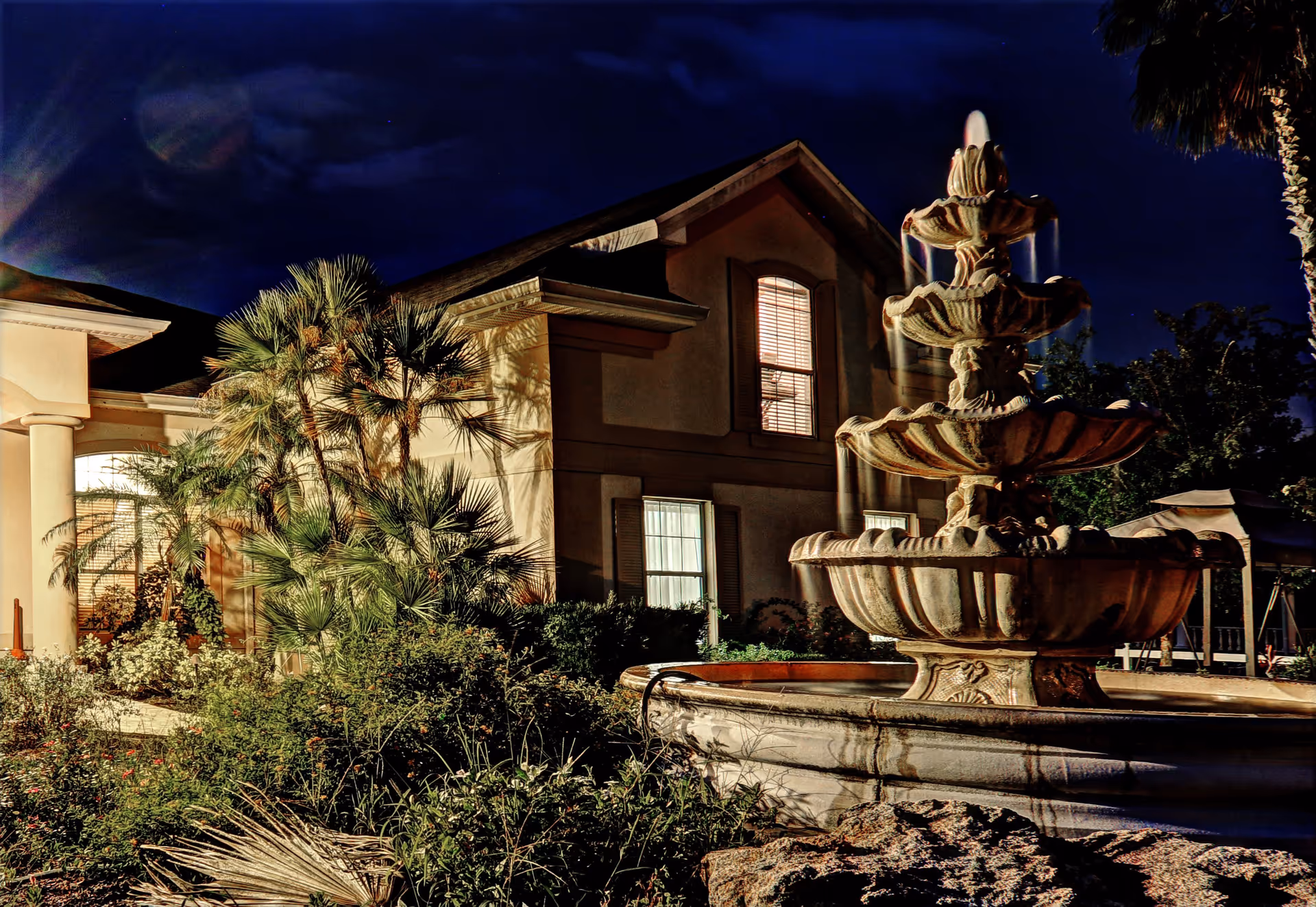 Night view of a building exterior with illuminated windows, surrounded by lush greenery and palm trees. In the foreground, there is a large multi-tiered stone fountain with water flowing from the top tier. The sky is dark blue with some clouds visible.