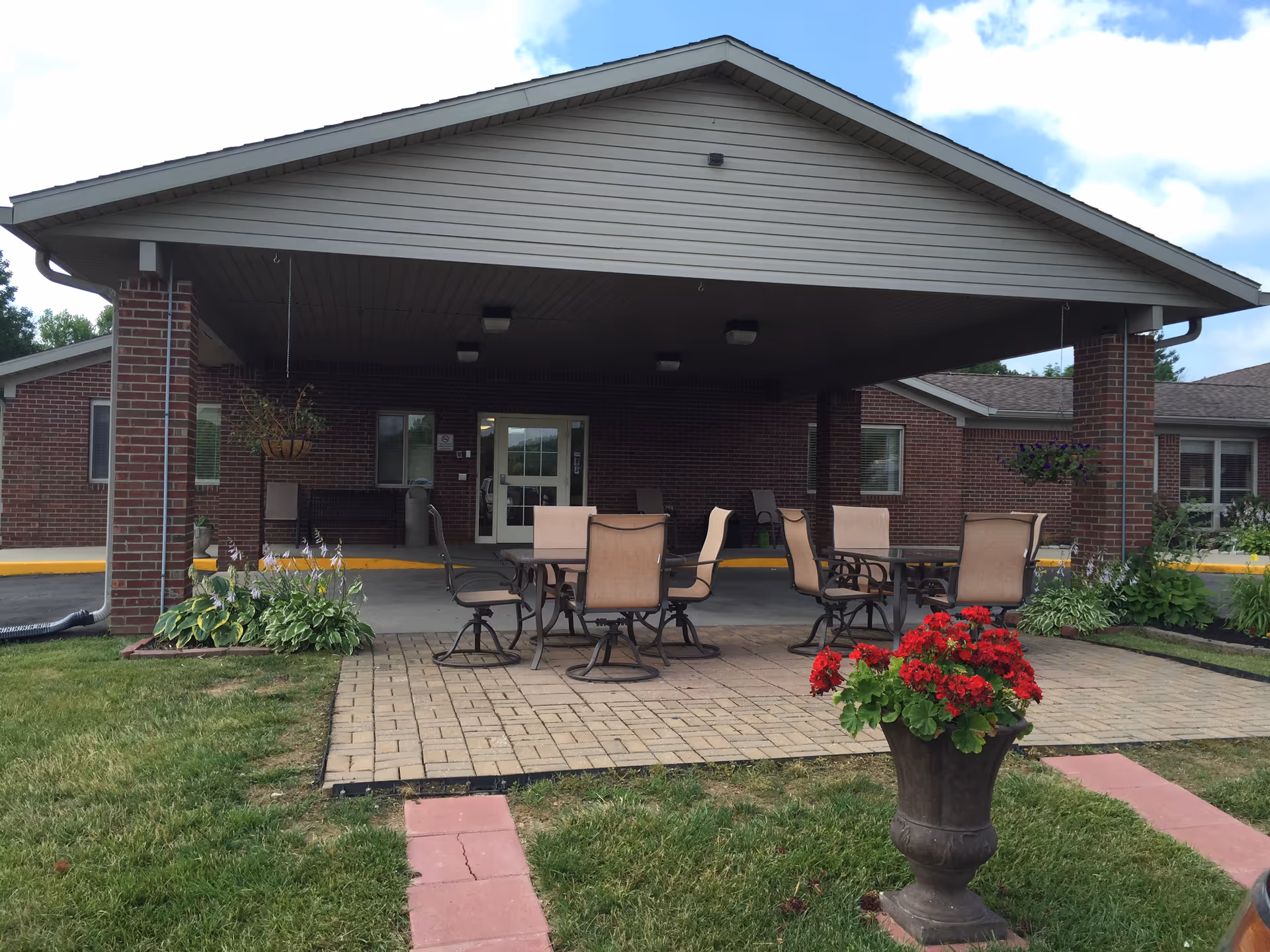 Covered entrance of a brick senior living facility with outdoor patio tables and chairs and a flower planter in front.