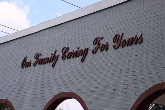 A close-up view of a white brick wall with red cursive text that reads 'Our Family Caring For Yours'. The top of the wall has a white trim and there are three red brick archways visible at the bottom. The sky is partly cloudy with some power lines visible above the wall.