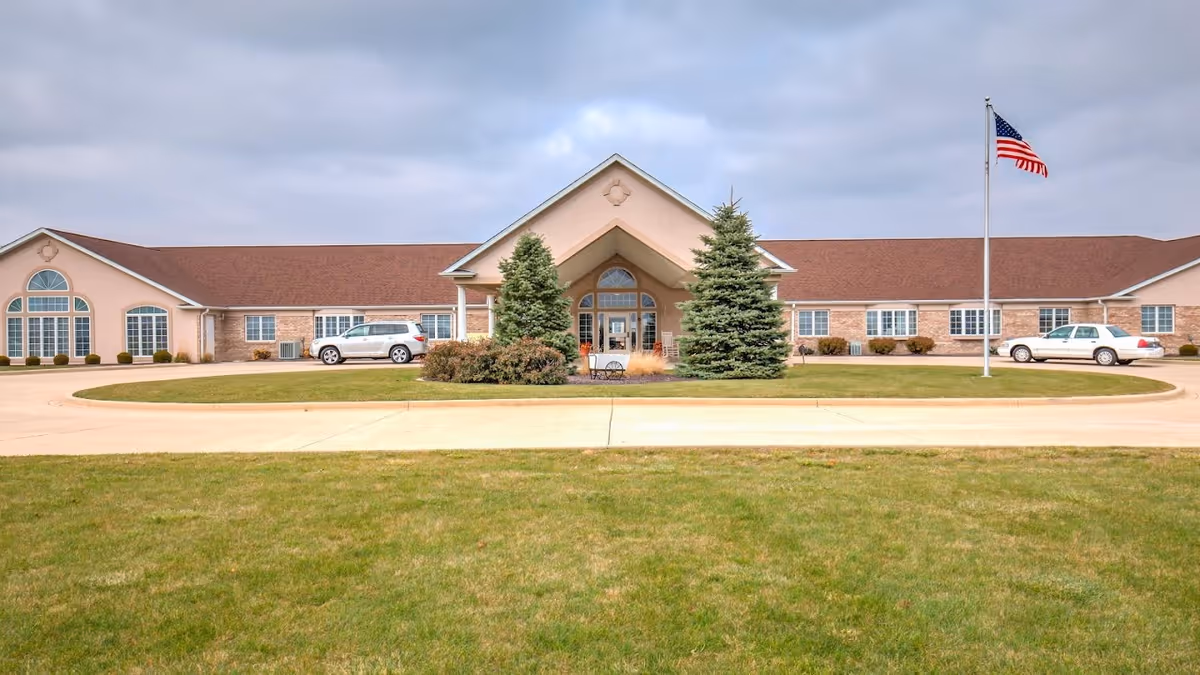 Front exterior view of Brookstone Estates of Tuscola, a single-story building with a peaked entrance, two evergreen trees, an American flag on a flagpole, and two cars parked near the entrance. The sky is cloudy and there is a large grassy area in the foreground.