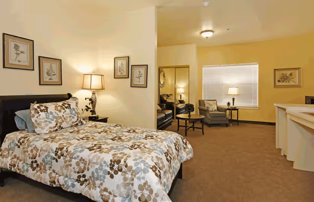 A spacious bedroom with a bed covered in a floral patterned comforter and pillows. There is a nightstand with a lamp and framed botanical prints on the wall. In the background, there is a sitting area with a chair, a small table with a lamp, and a mirrored closet door. The room has beige walls and carpeted floor.