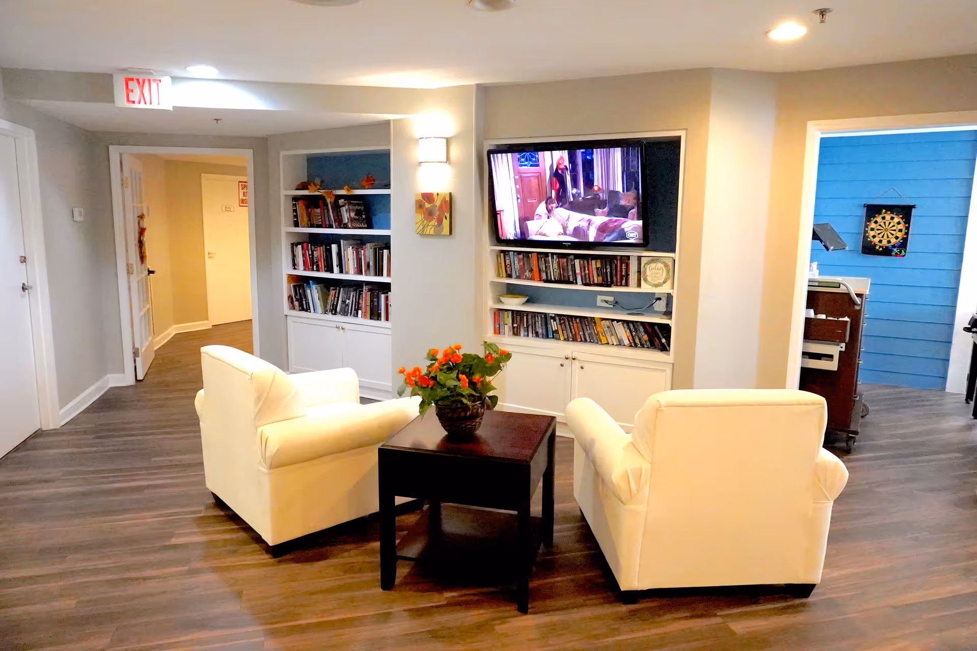 A cozy common area with two cream-colored armchairs facing a dark wooden table with a potted plant. Behind the chairs is a built-in bookshelf filled with books and a wall-mounted TV showing a scene from a show. The room has wood flooring, light-colored walls, and an exit sign above a doorway on the left. Another doorway on the right reveals a blue wall with a dartboard.