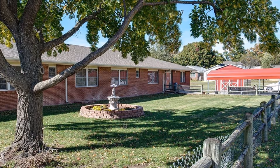 Front yard of a single-story brick building with a large tree, a circular flowerbed with a fountain, and a fenced grassy lawn.