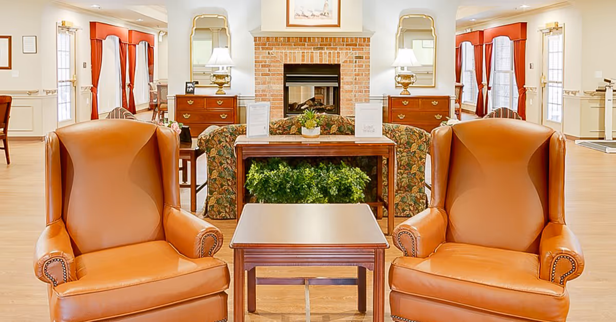 A cozy senior living facility common area featuring two brown leather armchairs facing a wooden coffee table. Behind the table is a floral-patterned sofa and a wooden console table with green plants underneath. The room has a brick fireplace centered on the back wall, flanked by two wooden dressers with table lamps and mirrors above them. The space is well-lit with natural light coming through windows with red curtains.