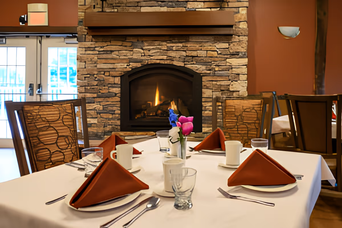 A dining table set for four with white tablecloth, brown folded napkins, white cups, glasses, and silverware in front of a stone fireplace with a fire burning inside. The room has wooden chairs with patterned cushions and a warm, cozy ambiance.