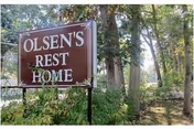 Brown sign reading "Olsen's Rest Home" surrounded by trees and foliage at the property entrance.