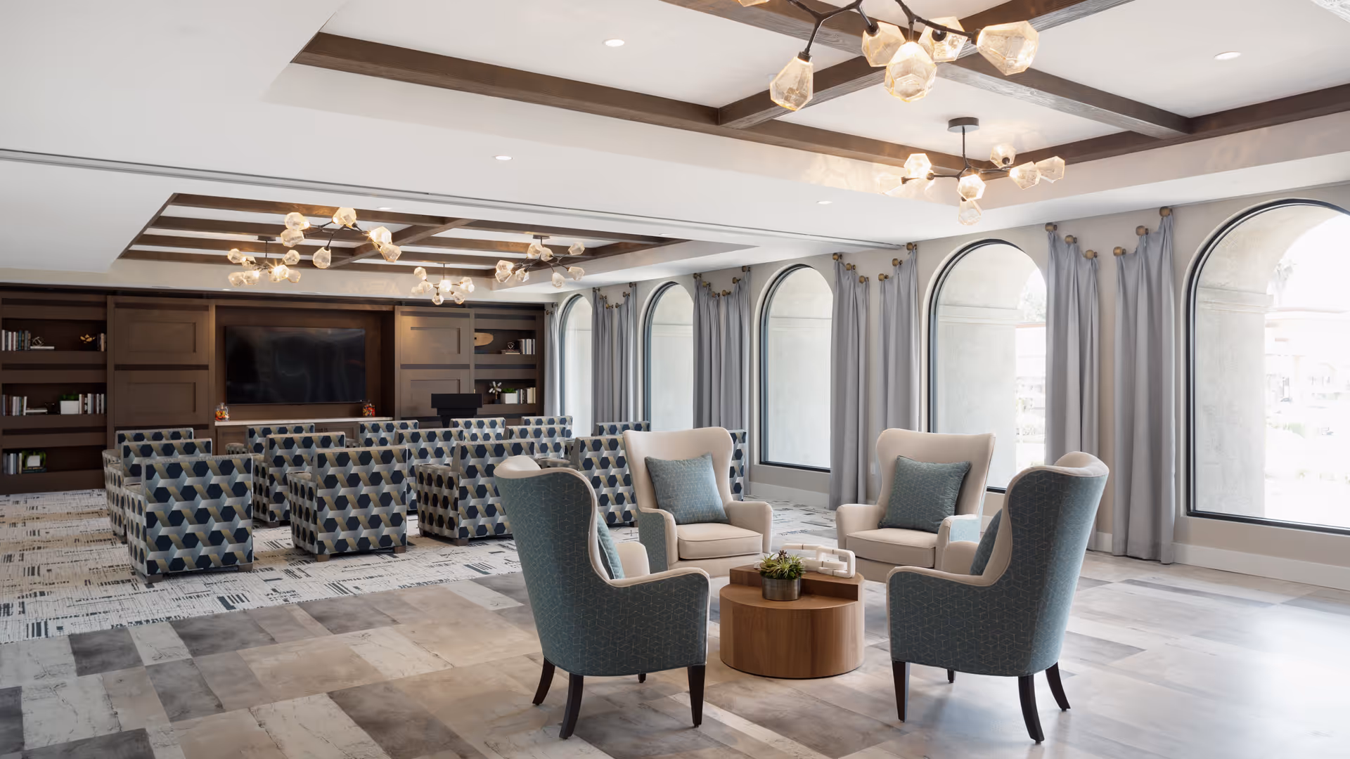 Bright, spacious senior living lounge with armchairs arranged around a round coffee table, patterned seating rows, arched windows, and a TV in built-in wooden shelving.