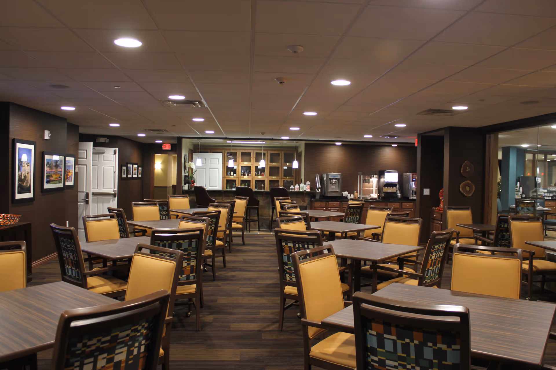 Interior view of a dining area in a retirement community with multiple tables and chairs arranged neatly. The chairs have yellow cushions and patterned backs. The room has warm lighting with ceiling lights and pendant lights over a counter area in the background. There are framed pictures on the walls and a beverage station with coffee machines and dispensers.
