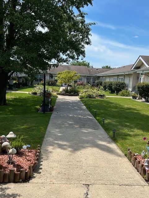 A concrete pathway leading through a well-maintained garden area with green grass, flower beds bordered by wooden logs, and small garden lights. There are trees and shrubs along the path, and a single-story building with multiple windows is visible in the background under a partly cloudy blue sky.
