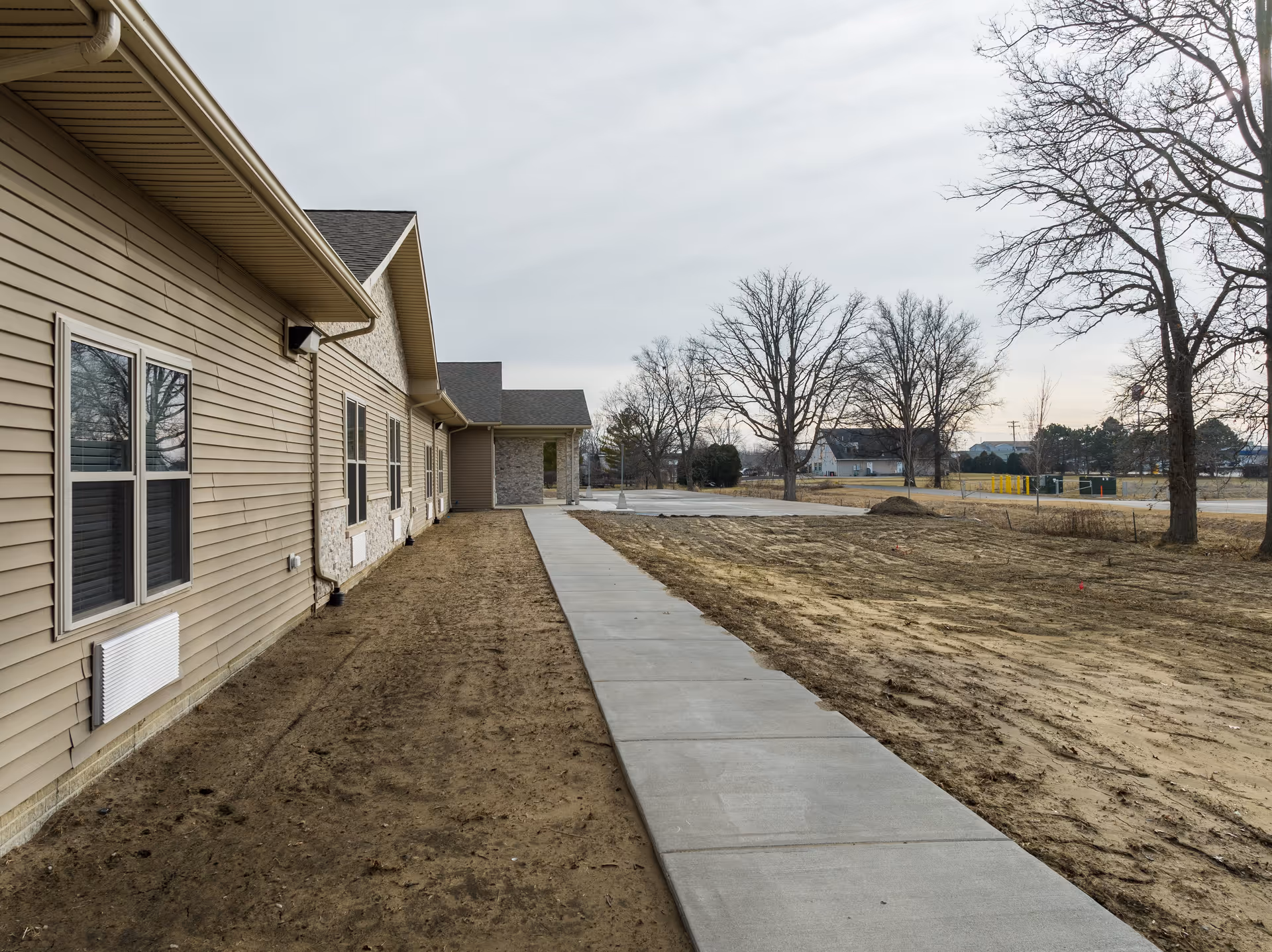 Side view of a building with beige siding and multiple windows, a concrete sidewalk running alongside it, bare soil on both sides of the sidewalk, leafless trees, and a cloudy sky in the background.