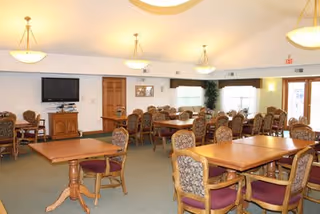 A spacious dining room with multiple wooden tables and cushioned chairs arranged neatly. The room has beige walls, carpeted floor, and ceiling lights. There is a flat-screen TV mounted on the wall and a wooden cabinet beneath it. Large windows with valances allow natural light to enter the room.