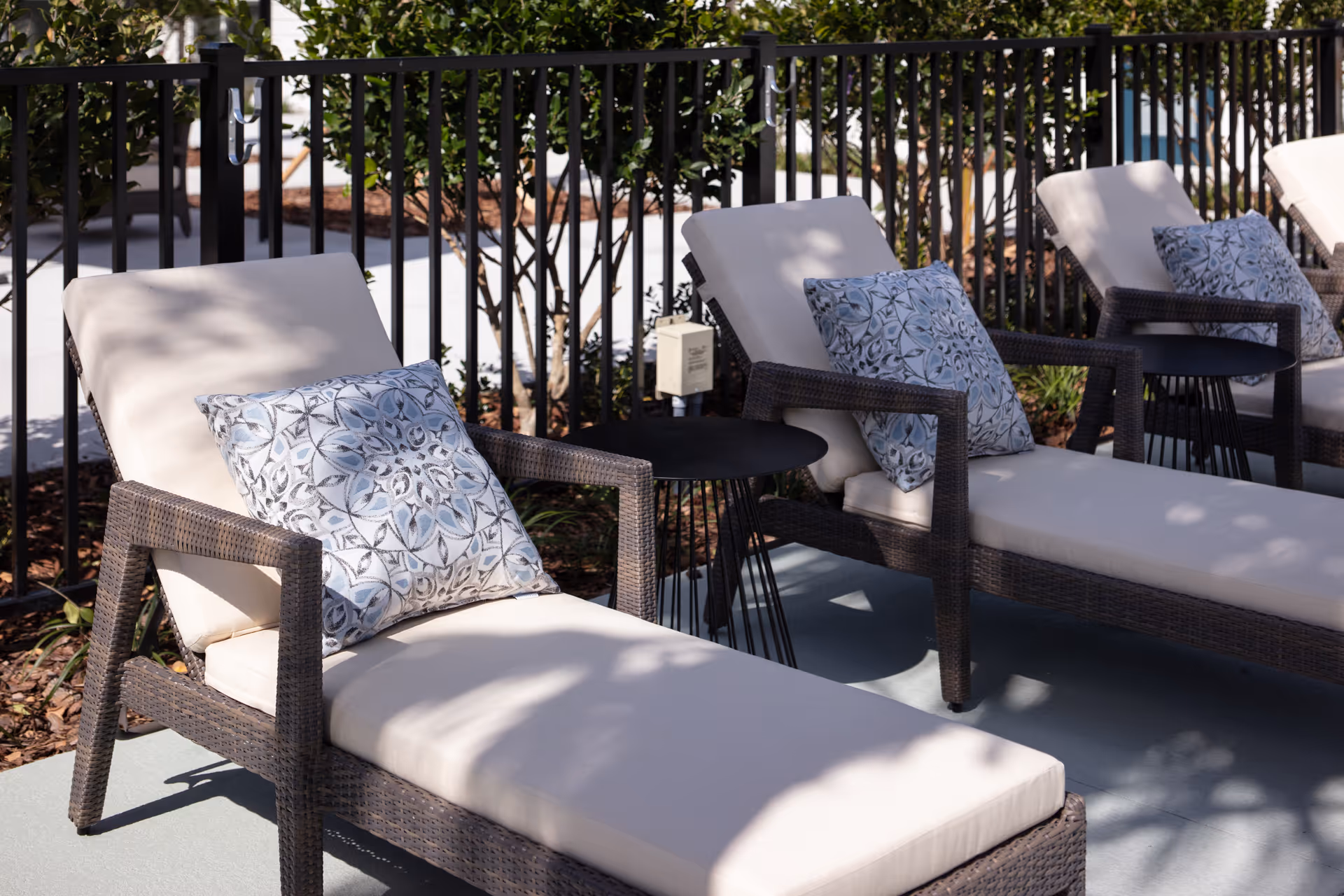 Outdoor patio area with several cushioned lounge chairs featuring patterned pillows, small black side tables between the chairs, and a black metal fence with greenery in the background.