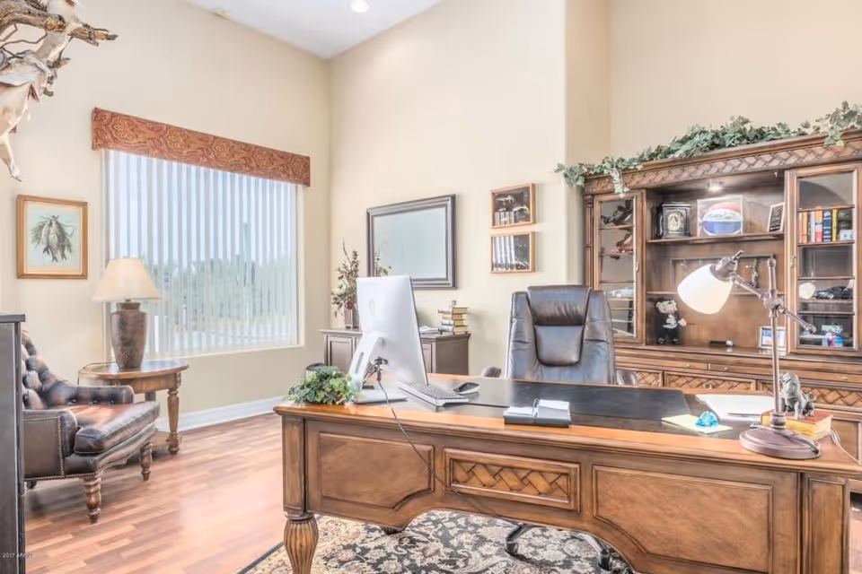 Sunlit furnished office with a wooden desk, leather chair, bookcase, and a large window with vertical blinds.