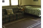 A cozy room with a plaid sofa on the left and a single bed with a green patterned quilt on the right. There is a small wooden nightstand between the sofa and bed with a lamp and some items on it. A window with curtains is behind the sofa, and a framed picture hangs on the wall above the bed. The floor is carpeted with a rectangular area rug featuring a simple border design.