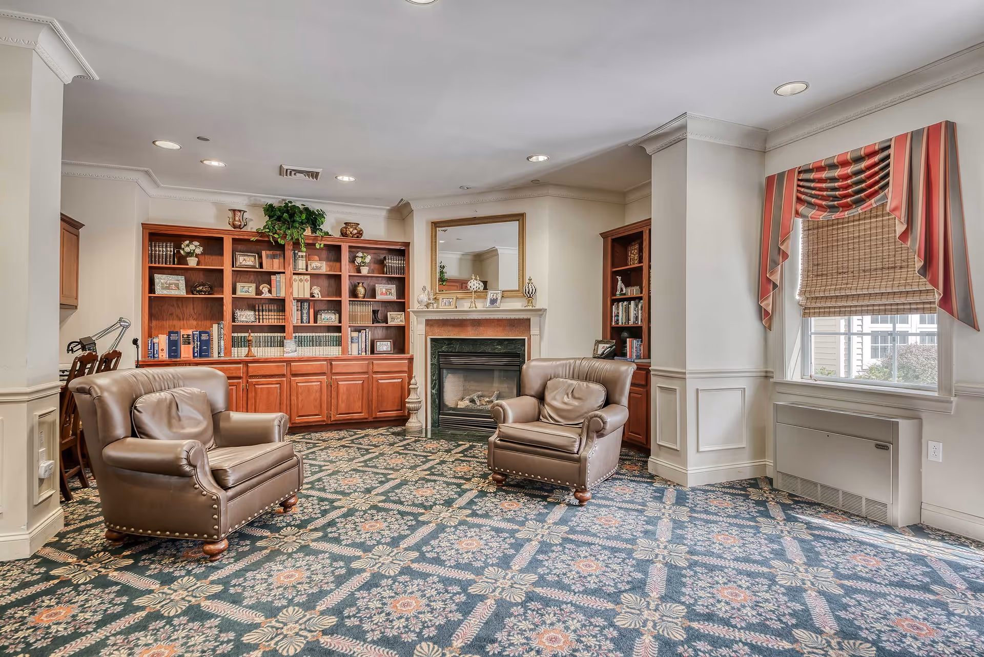 A cozy lounge with leather armchairs, built-in wooden bookshelves, a fireplace, and a patterned carpet near a window with a valance.