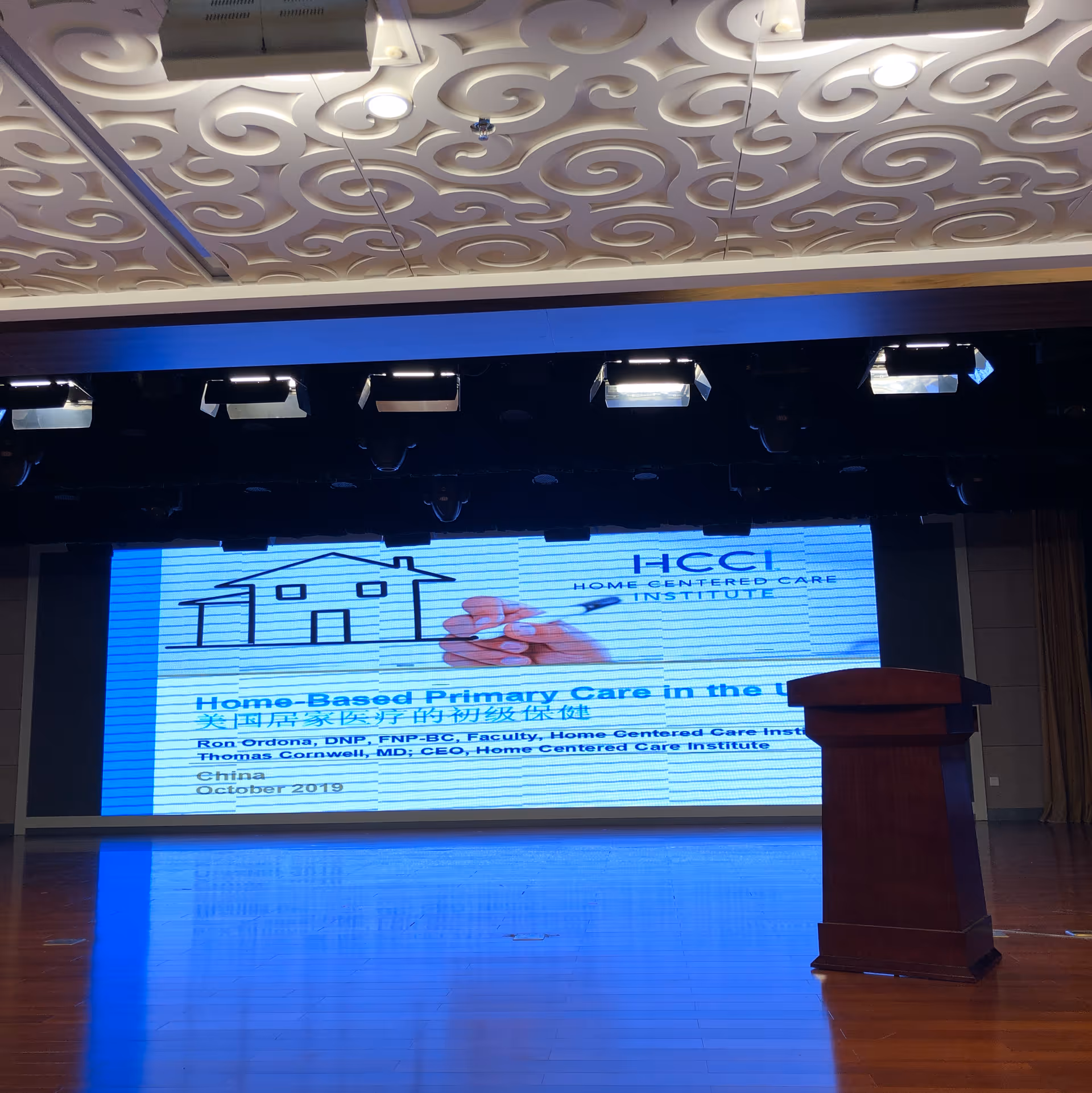 Stage and podium in an empty auditorium with a large illuminated presentation screen showing a slide about home-based primary care.