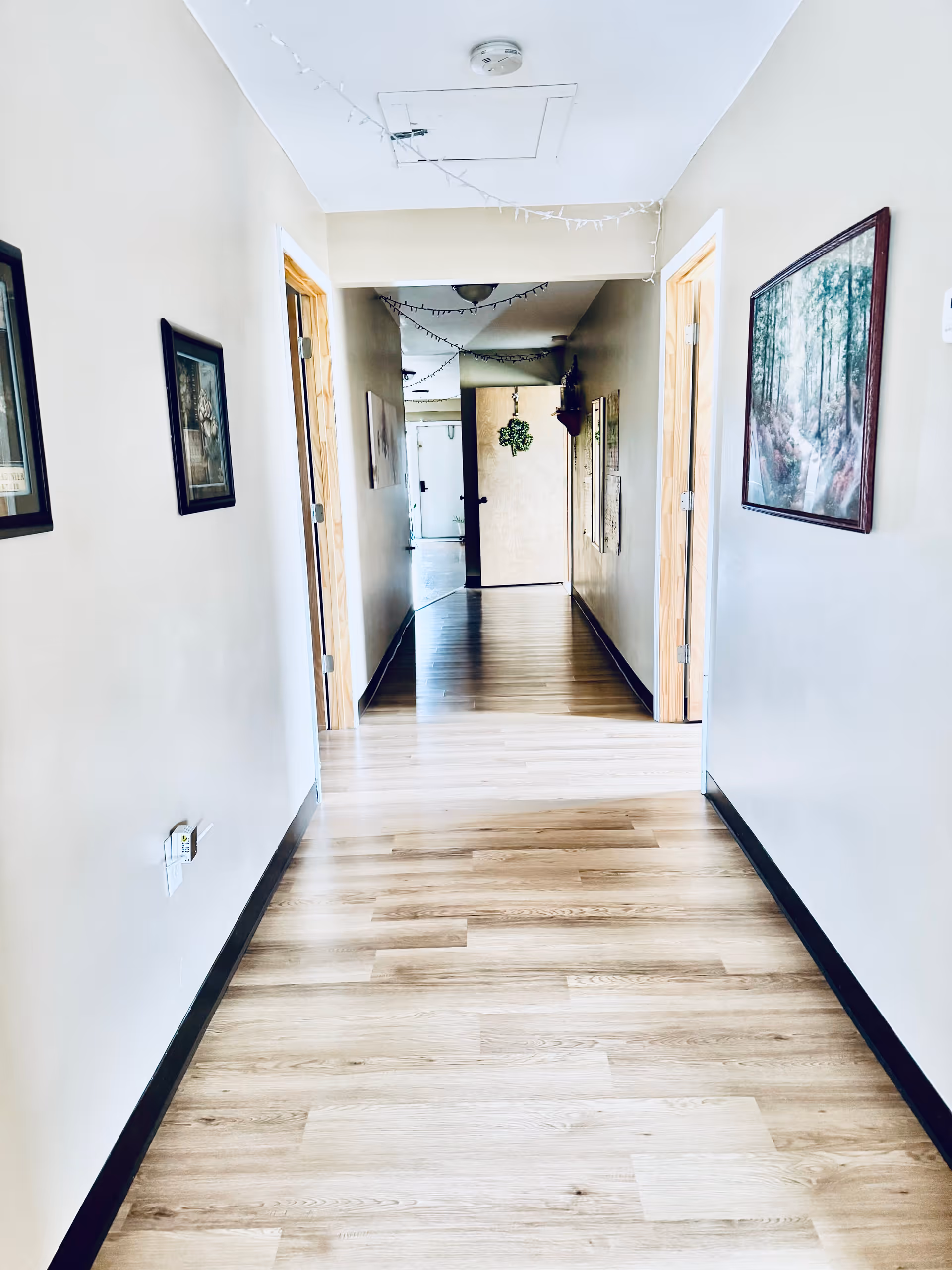 A bright hallway with light wood flooring and beige walls. There are framed pictures hanging on both sides of the walls. The hallway has several doorways leading to other rooms and a door at the far end with a green wreath decoration. String lights are hung along the ceiling.
