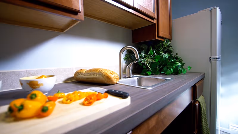 A kitchen countertop with a cutting board holding sliced yellow and orange bell peppers and a knife. There is a loaf of bread and a bowl on the counter near a stainless steel sink with a faucet. Wooden cabinets are above the counter, and a refrigerator is visible in the background with a green plant next to the sink.