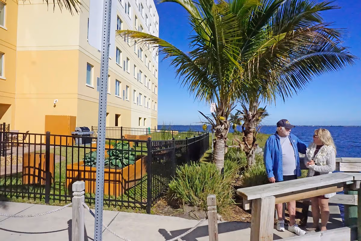 Two people standing and talking near a wooden picnic table by a waterfront with palm trees and a multi-story building in the background under a clear blue sky.