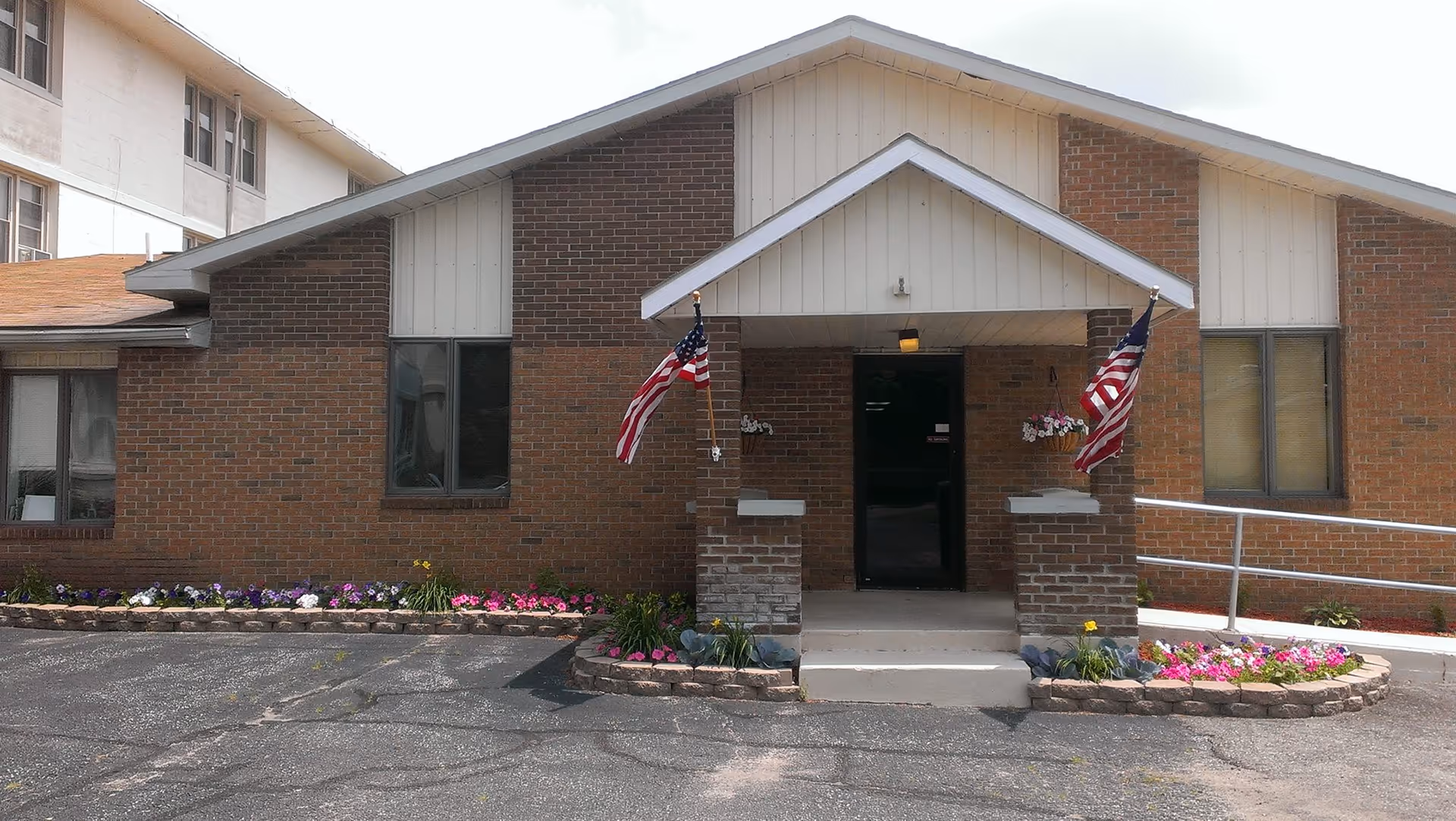 Front exterior view of a single-story brick building with a peaked roof entrance. Two American flags are mounted on either side of the entrance, which has a small porch with steps and flower beds with colorful flowers in front. There is a wheelchair ramp on the right side of the entrance.