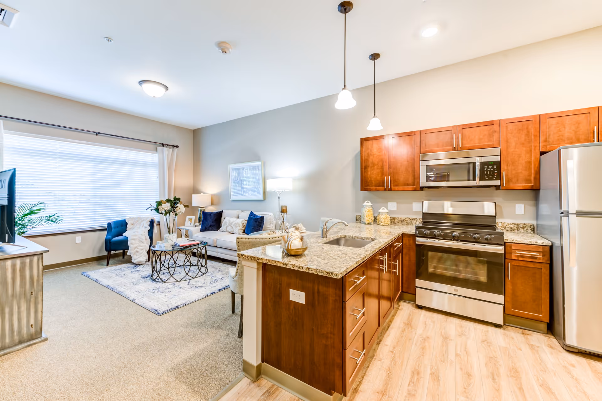 Open-plan apartment interior with a modern kitchen featuring granite counters and stainless appliances next to a furnished living room with a sofa and large window.