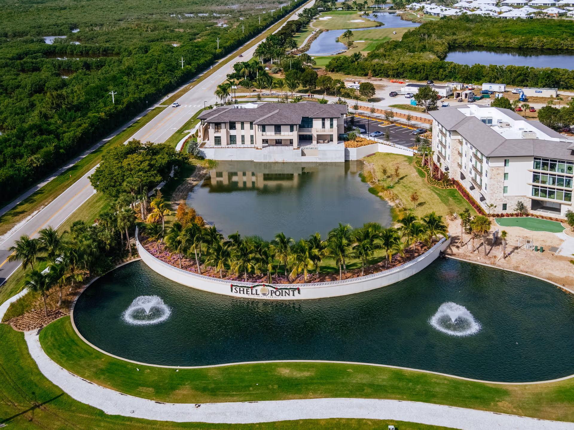 Aerial view of Shell Point Welcome Center showing a landscaped area with two water fountains in a pond, palm trees, a curved wall with the Shell Point sign, and multiple buildings surrounded by greenery and roads.