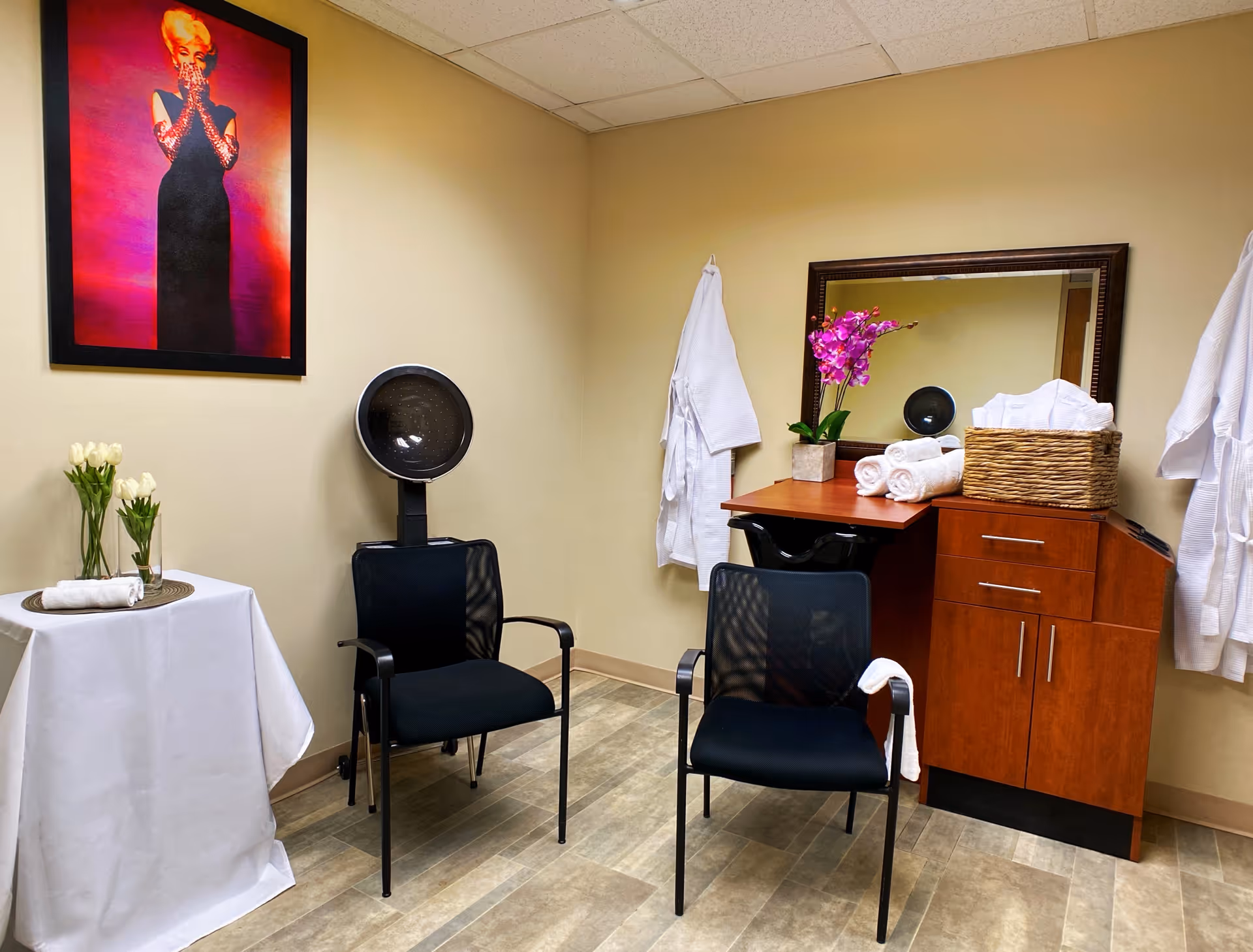 Interior of a salon or spa room with two black chairs, a hair dryer, a wooden cabinet with a mirror, rolled white towels, a basket, a purple orchid plant, and white robes hanging on the wall. A small round table with a white tablecloth holds two vases with white flowers and a rolled towel. A framed colorful portrait of a woman in a black dress is on the wall.