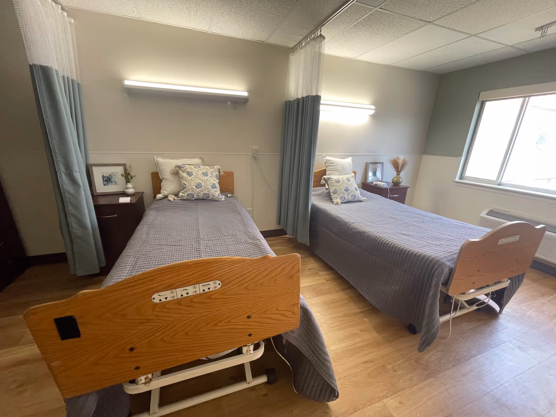 A room with two single hospital-style beds separated by a privacy curtain. Each bed has a wooden headboard and footboard, gray bedspreads, and decorative pillows. There are nightstands next to each bed with framed pictures and small decorative items. The room has wood flooring, a window letting in natural light, and wall-mounted lights above each bed.