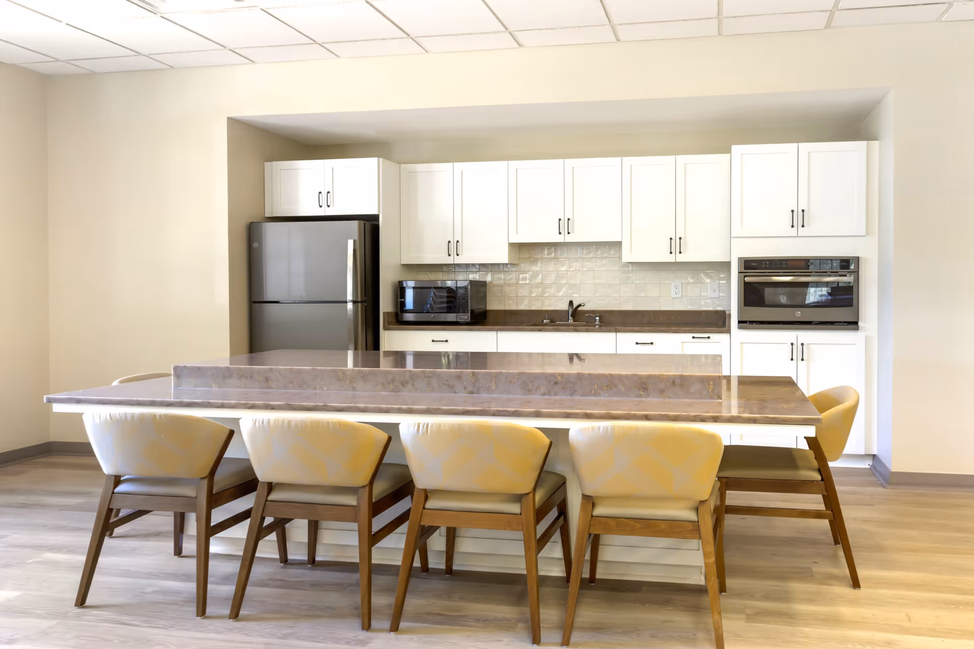 Bright communal kitchen with a large central island surrounded by six chairs, white cabinets, and stainless appliances.