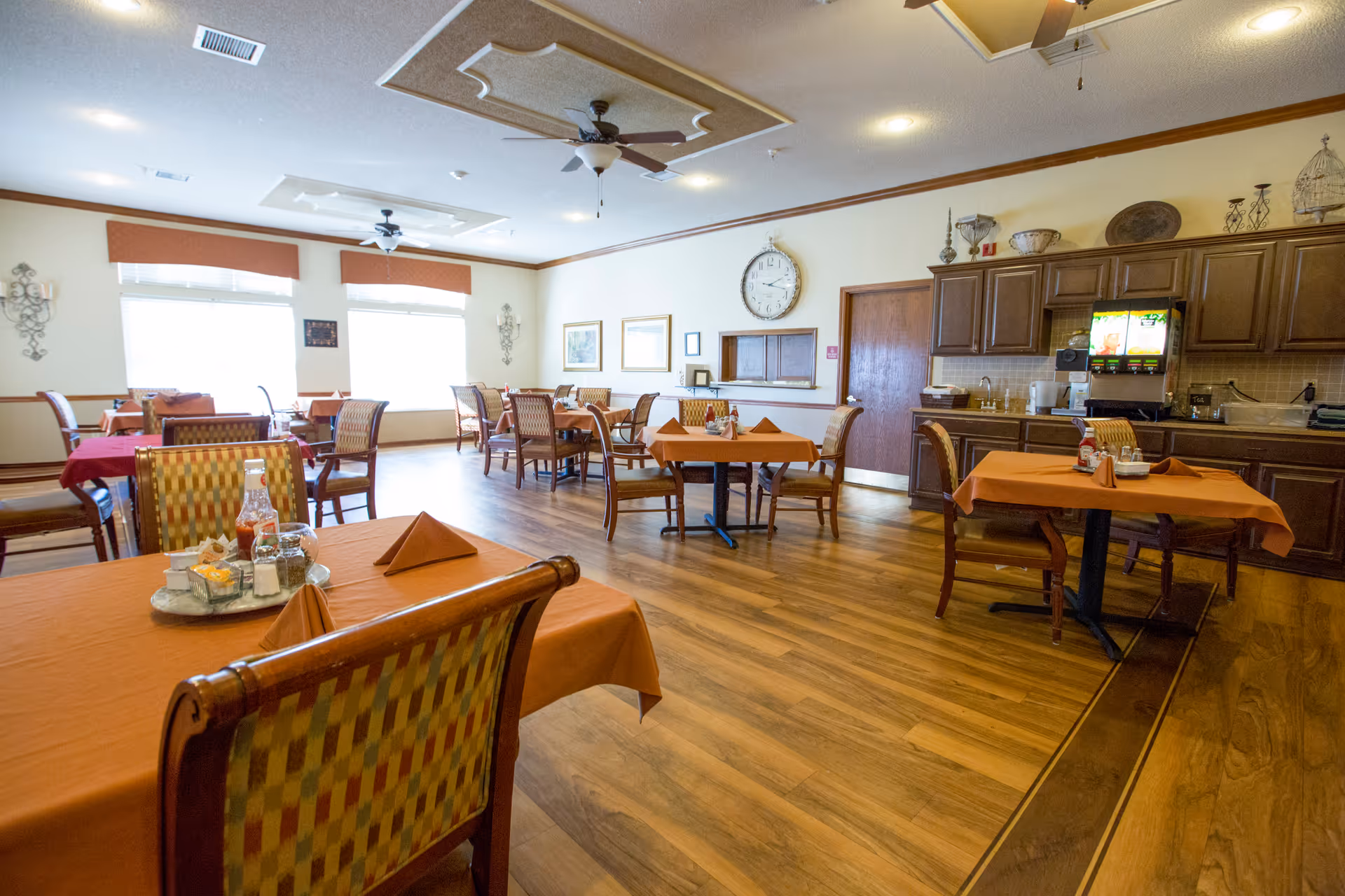 A dining room with several tables covered in orange and red tablecloths, each set with folded napkins and condiments. The room has wooden flooring, large windows with valances, ceiling fans, and a wall clock. There is a counter area with cabinets and beverage dispensers on the right side.