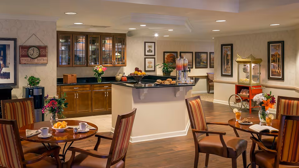 A cozy dining area in a senior living facility featuring round wooden tables with striped cushioned chairs. On the tables are cups, plates with food, flowers in vases, and glasses of iced tea. In the background, there is a counter with a beverage dispenser, a fruit bowl, and baked goods. Wooden cabinets with glass doors are mounted on the wall, and framed artwork decorates the walls. A vintage-style popcorn machine is visible on the right side of the room.