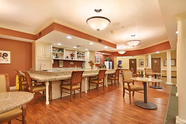 A spacious dining area with wooden floors and warm-toned walls. The room features a long granite countertop with several wooden chairs lined up along it. There are round tables with chairs scattered throughout the room. The ceiling has recessed lighting and hanging light fixtures. The back wall has built-in shelves and cabinets, and there are framed pictures and plants decorating the space.