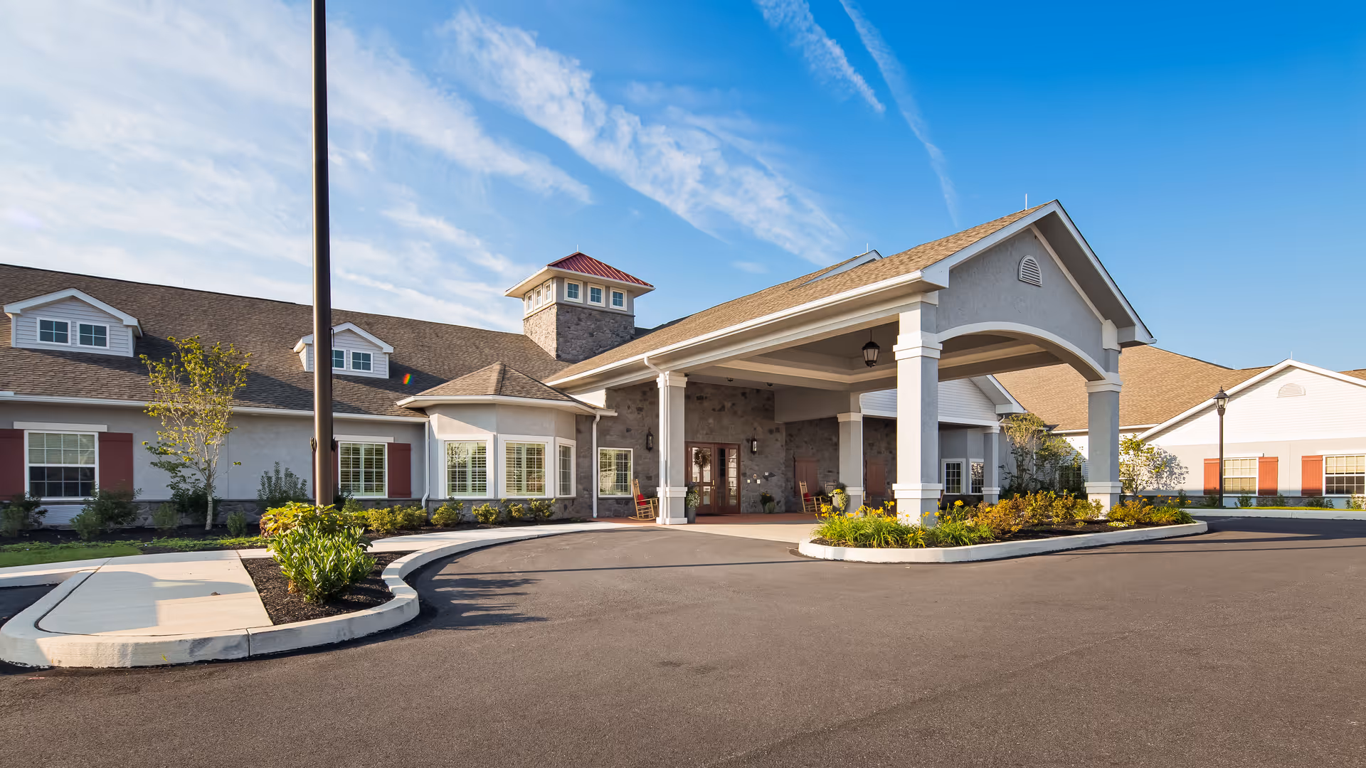 Exterior view of a senior living facility named Legend of Lititz showing the main entrance with a covered drop-off area, landscaped greenery, and a clear blue sky.