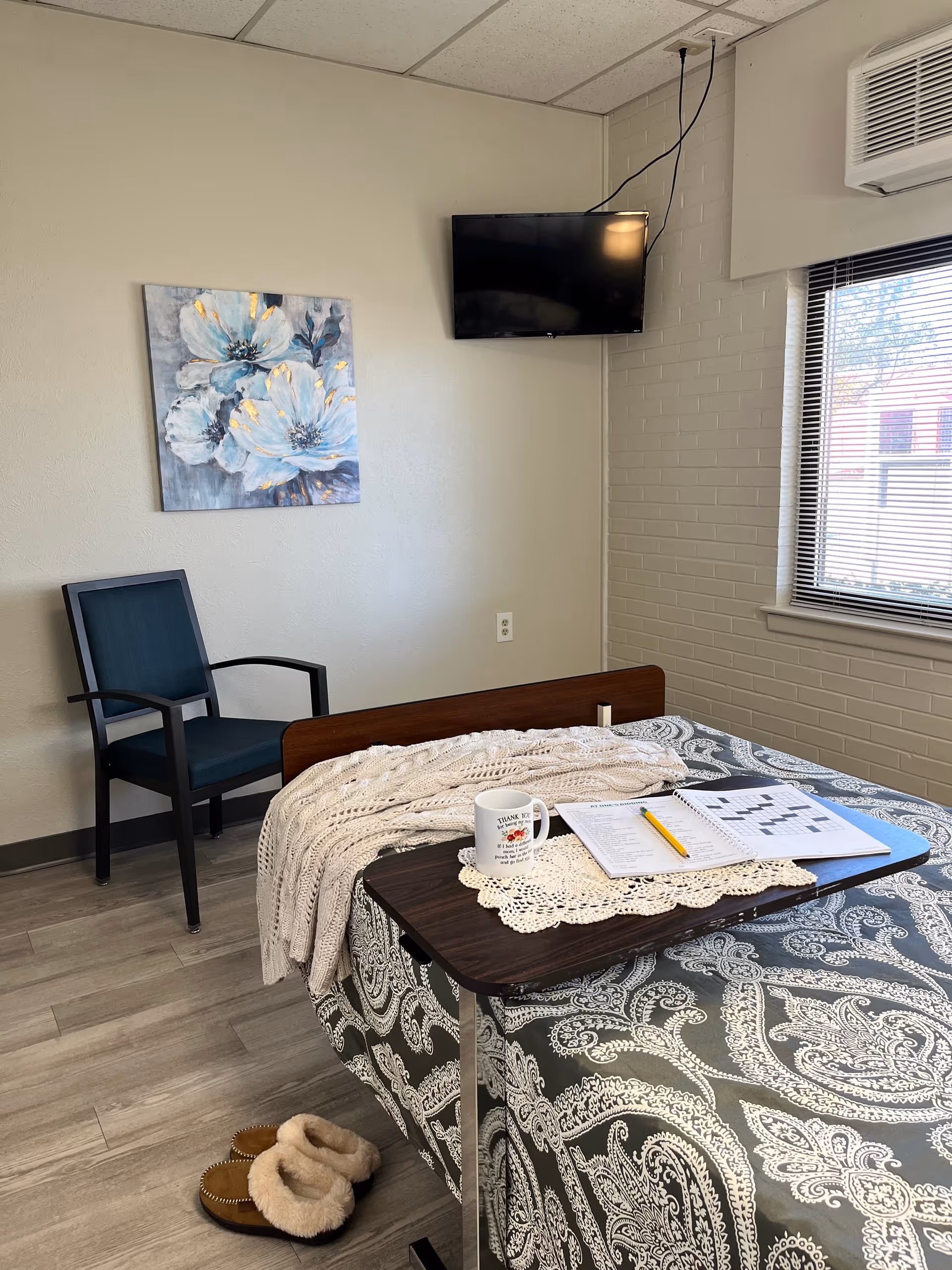 A nursing facility room with a bed covered in a patterned bedspread and a knitted blanket. On the bed is a tray table holding a crossword puzzle book, a pencil, and a coffee mug. Next to the bed on the floor are a pair of slippers. A blue chair is positioned against the wall, which has a painting of white flowers. A flat-screen TV is mounted on the wall near a window with blinds, and an air conditioning unit is above the window.