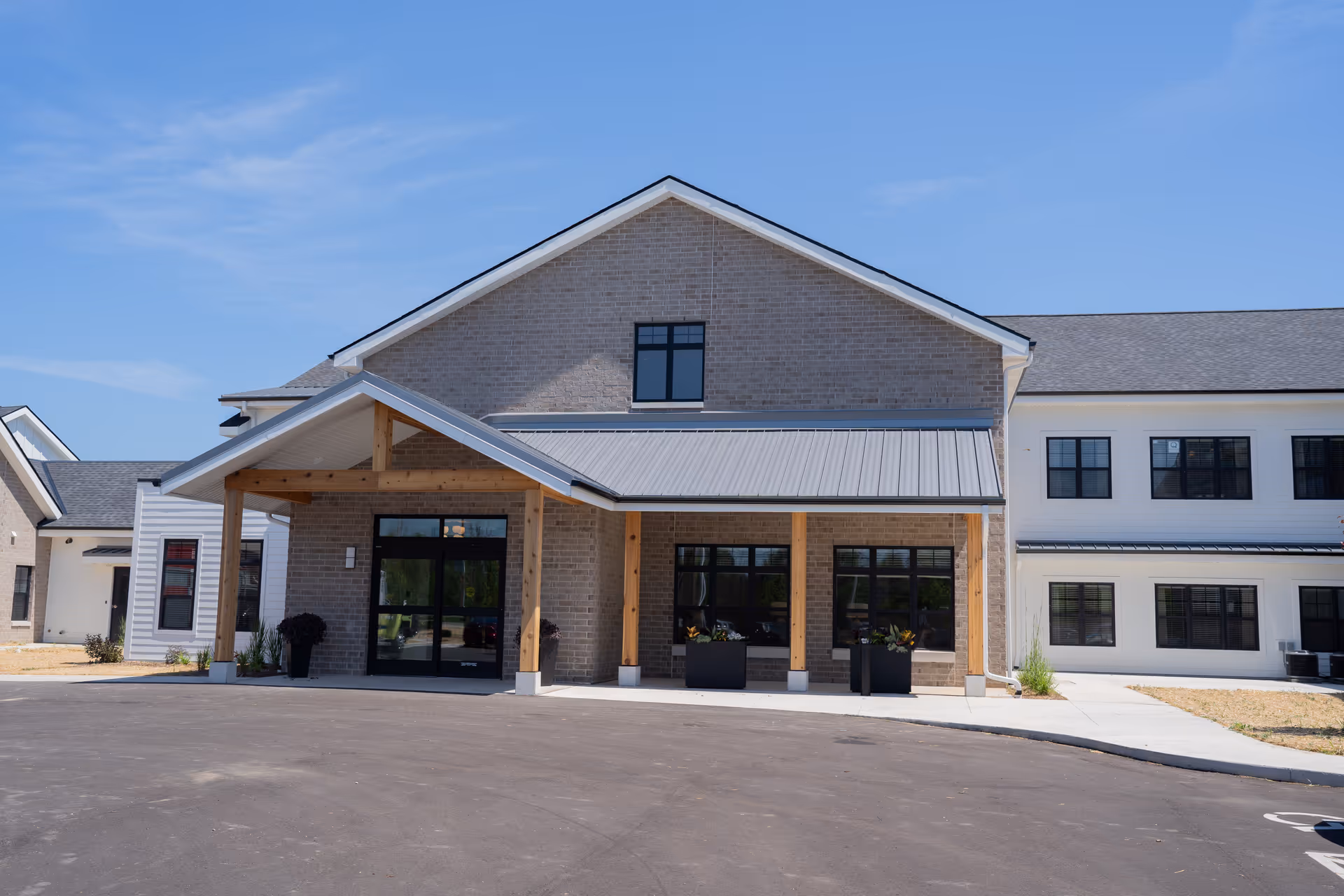Front exterior view of Lumia Mequon Assisted Living & Memory Care building with a covered entrance supported by wooden beams, large windows, and a paved driveway under a clear blue sky.