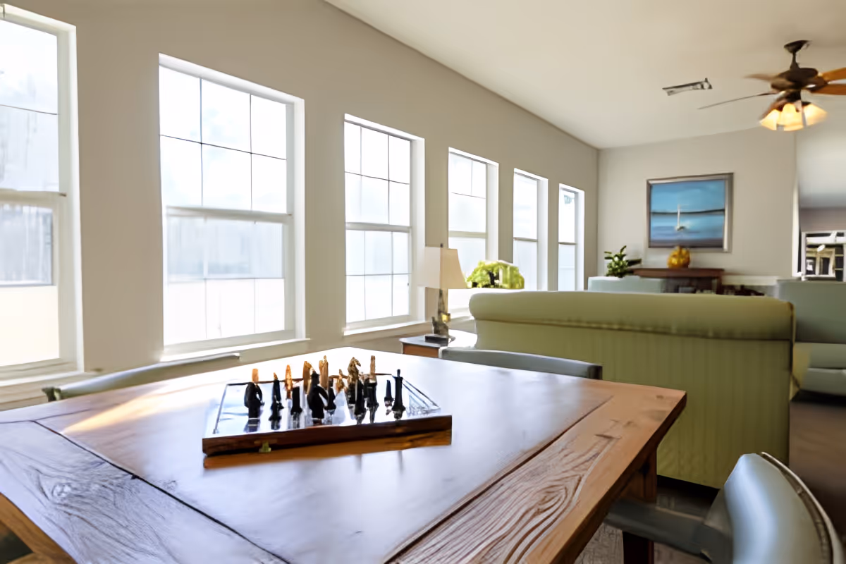 Bright communal living room with a wooden table and chessboard in the foreground, rows of tall windows and green sofas in the background.
