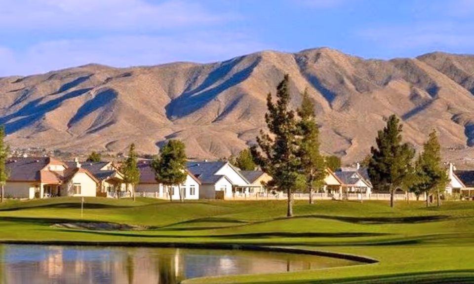Manicured golf-course greens and a pond in front of single-story homes with trees and mountains in the background.