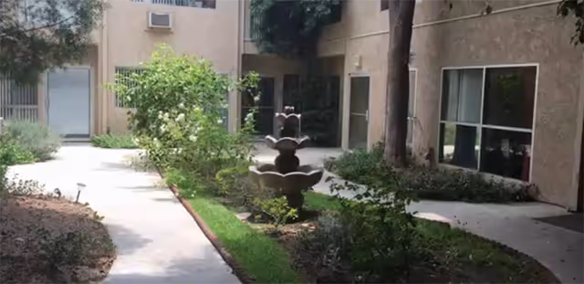 Sunlit courtyard with a three-tiered fountain, walkways, shrubs, and surrounding apartment windows and doors.