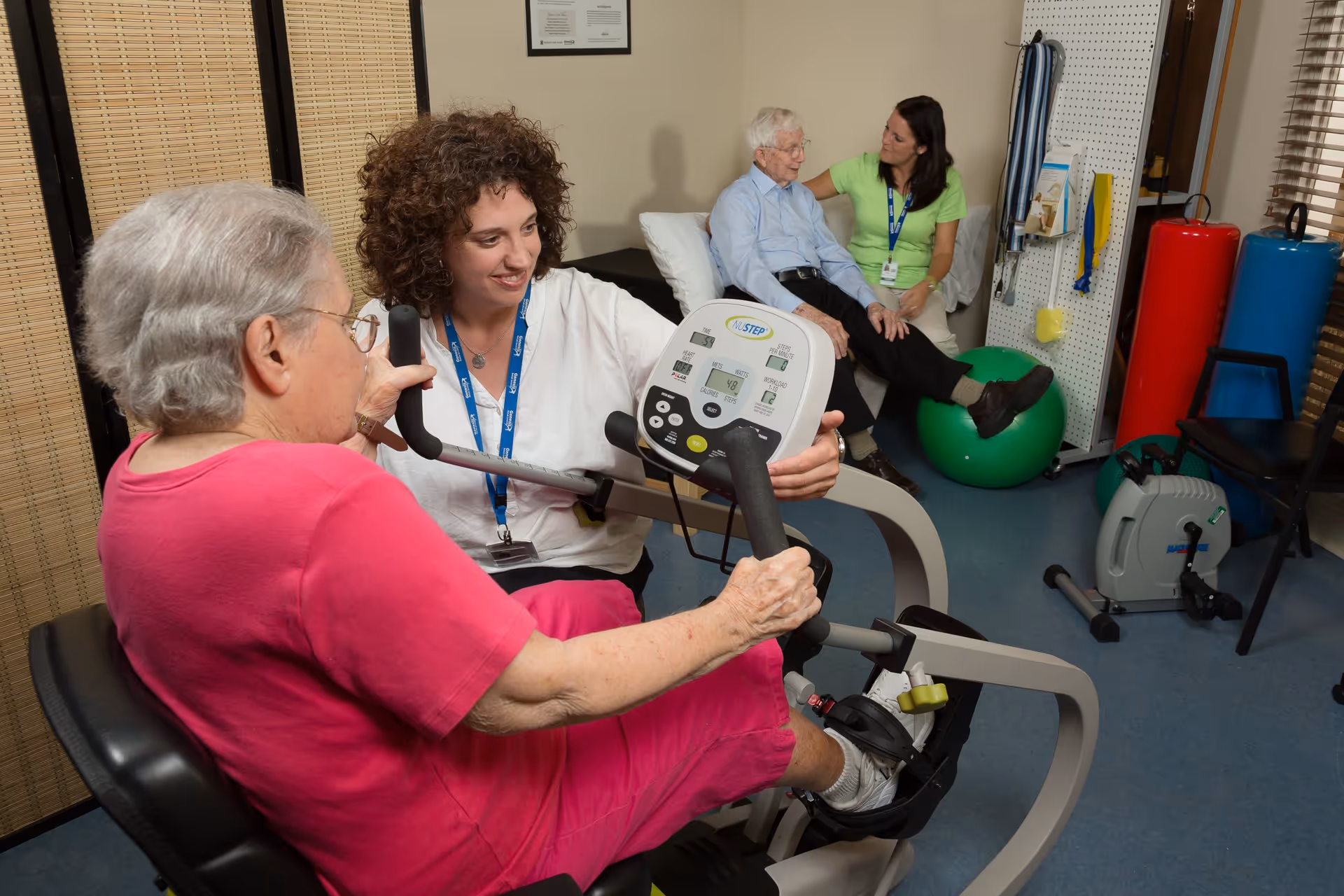 An elderly woman in a pink outfit uses a seated exercise machine while a female staff member in a white shirt and blue lanyard assists her. In the background, an elderly man sits on a green exercise ball talking with another female staff member in a green shirt. The room contains various physical therapy and exercise equipment.